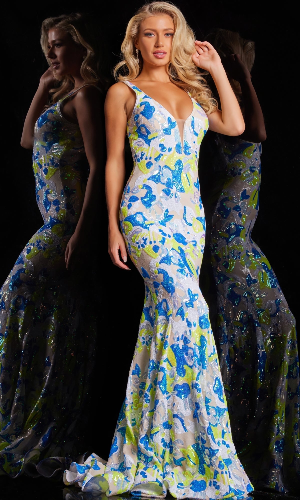 A woman stands beside her reflection in a fitted, floor-length Jovani V-Back Long Sequin-Print Prom Dress 26130 with abstract yellow and blue floral patterns, set against a black background.